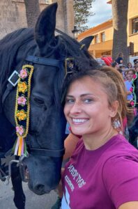 Kobieta w bordowej koszulce uśmiecha się do aparatu, przytulając ozdobionego czarnego konia podczas festiwalu