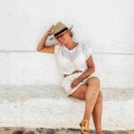 Kobieta w jasnym letnim stroju i słomkowym kapeluszu relaksuje się, siedząc na białej ławce w urokliwej uliczce Minorki.