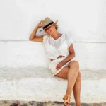 Kobieta w jasnym letnim stroju i słomkowym kapeluszu relaksuje się, siedząc na białej ławce w urokliwej uliczce Minorki.