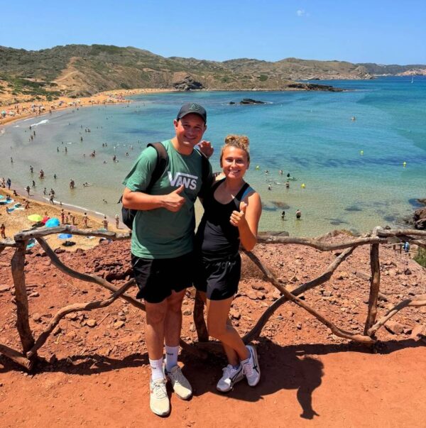 Uśmiechnięta para pozuje na tle turkusowej wody i złocistej plaży Cala Cavalleria na Minorce.