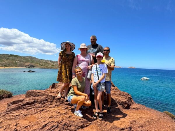 Grupa turystów pozuje na tle turkusowego morza na Minorce, stojąc na charakterystycznych czerwonych skałach w okolicy Cala Pregonda.