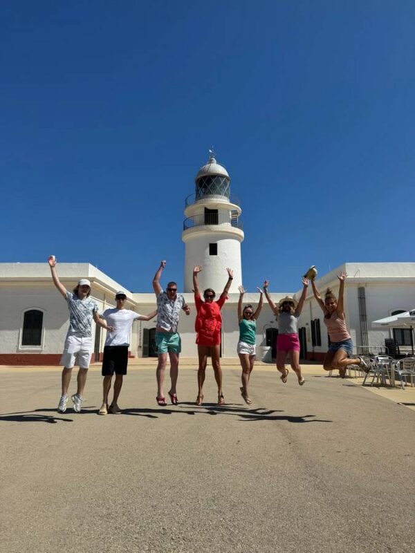 Grupa turystów skacząca w radosnym geście przed latarnią morską Faro de Cavalleria na Minorce. Słoneczna pogoda, błękitne niebo i biała architektura tworzą malowniczy krajobraz.