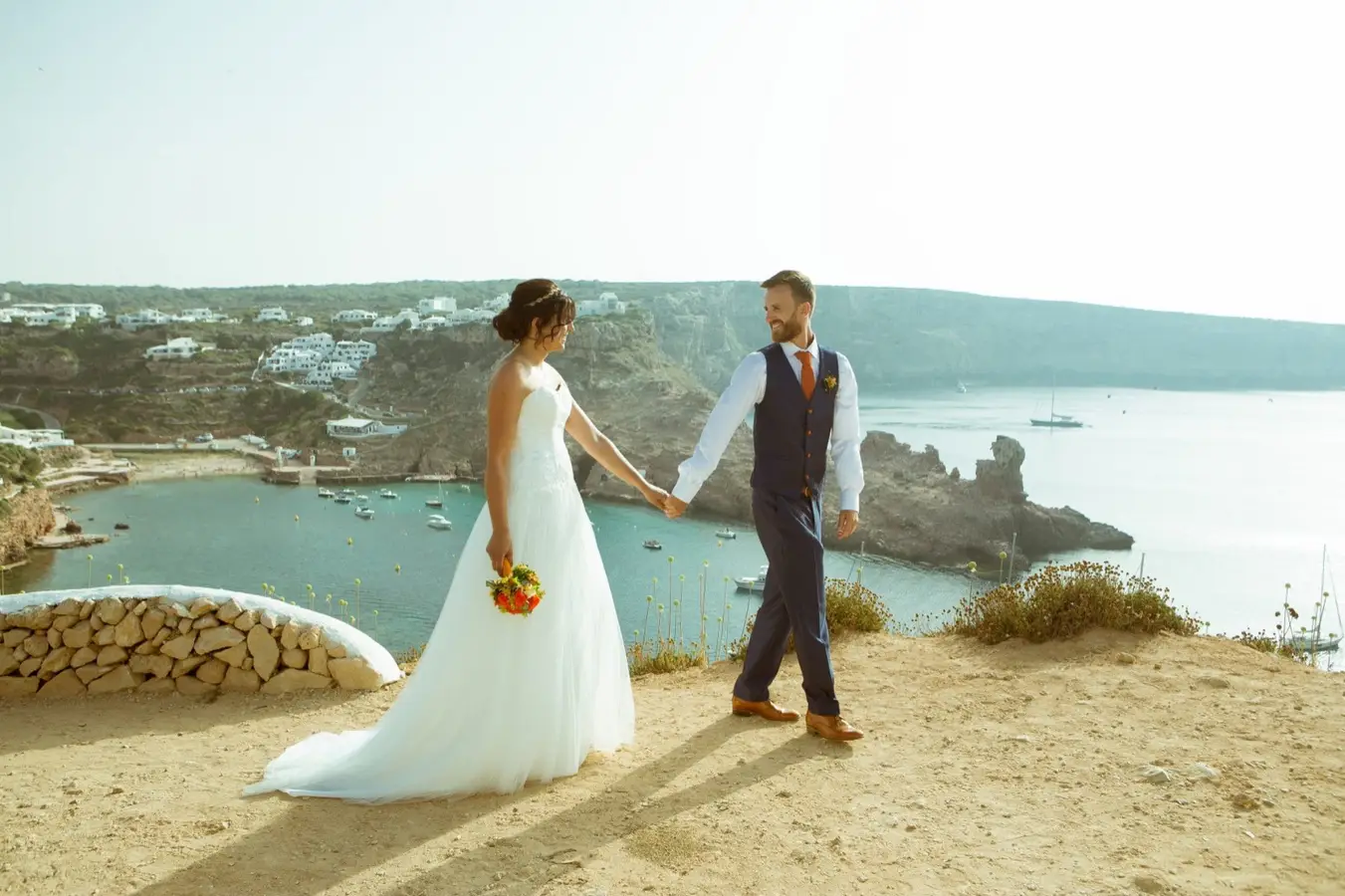 Para młoda trzymająca się za ręce na klifie z widokiem na zatokę na Minorce. Panna młoda w białej sukni z bukietem kwiatów, pan młody w eleganckim garniturze z kamizelką. W tle białe domy na wzgórzu, turkusowa woda i zacumowane łódki. Romantyczna sceneria śródziemnomorskiego wybrzeża.