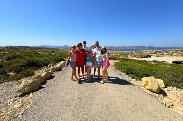 Grupa turystów na malowniczym szlaku w rezerwacie przyrody na Minorce. Widok na surowy krajobraz wyspy, porośnięty śródziemnomorską roślinnością, z górami i błękitnym morzem w tle.