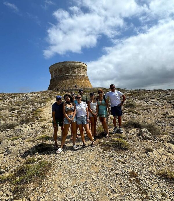 Grupa turystów na Minorce podczas wycieczki do Torre de Fornells, zabytkowej wieży obronnej na wzgórzu, otoczonej kamienistym krajobrazem.