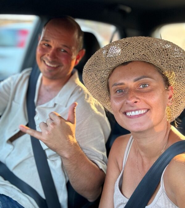 Uśmiechnięta kobieta w słomkowym kapeluszu i mężczyzna w lnianej koszuli w samochodzie, gotowi na wakacyjną przygodę.