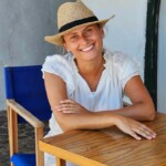 Uśmiechnięta kobieta w słomkowym kapeluszu odpoczywa przy drewnianym stoliku na Minorce, ciesząc się śródziemnomorską atmosferą.