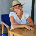 Uśmiechnięta kobieta w słomkowym kapeluszu odpoczywa przy drewnianym stoliku na Minorce, ciesząc się śródziemnomorską atmosferą.