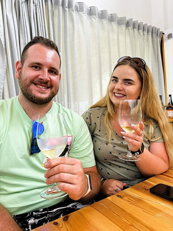 Uśmiechnięta para turystów siedzi przy drewnianym stole w winnicy na Minorce, trzymając kieliszki białego wina. Mężczyzna ma brodę, nosi jasną koszulkę i ma zawieszone na kołnierzyku okulary przeciwsłoneczne. Kobieta ma długie blond włosy, nosi okulary na głowie oraz oliwkową koszulkę z małymi wzorami. W tle widać zasłony i butelki wina na półce.