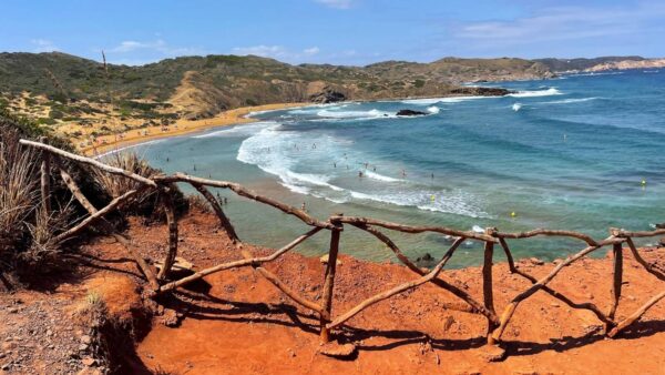 Widok na dziką plażę na Minorce z czerwonymi klifami i błękitnym morzem. W tle piaszczysta plaża otoczona zielonymi wzgórzami, a w wodzie kąpiący się ludzie. Na pierwszym planie drewniane ogrodzenie z gałęzi.