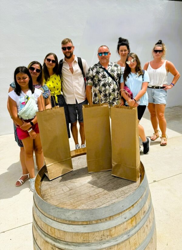 Grupa turystów pozuje przy drewnianej beczce w winnicy na Minorce. Przed nimi stoją trzy papierowe torby z zakupionymi winami. Ubrani w letnie stroje, z okularami przeciwsłonecznymi, wyglądają na zadowolonych z wizyty.