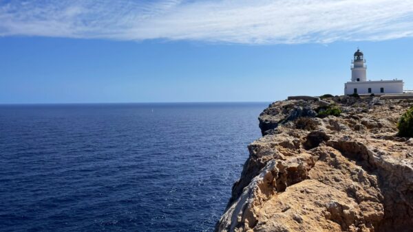 Biała latarnia morska na skraju skalistego klifu z widokiem na bezkresne, błękitne Morze Śródziemne. Na horyzoncie widać mały żagiel, a niebo zdobią delikatne chmury. Surowy krajobraz kontrastuje z spokojem otaczającej przestrzeni.