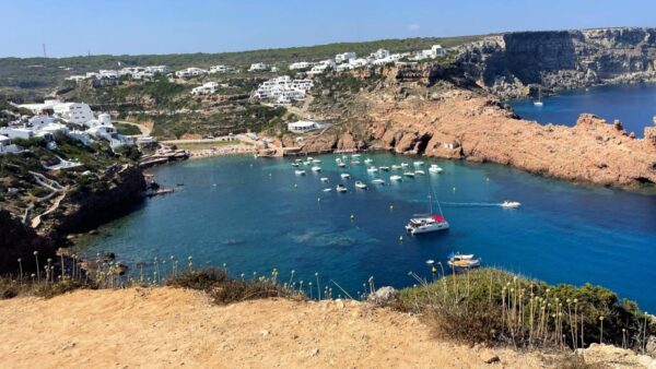 Malownicza zatoka otoczona skalistymi klifami i białymi śródziemnomorskimi domkami wtopionymi w zielone wzgórza. W spokojnej, turkusowej wodzie unoszą się liczne łodzie, a na piaszczystej plaży widać plażowiczów cieszących się słońcem. Idealne miejsce na relaks z widokiem na naturalne piękno Minorki.