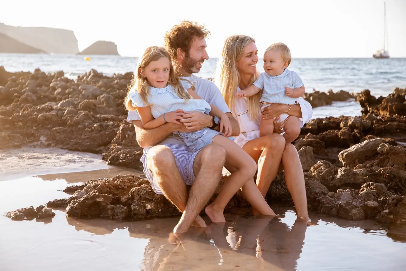 Szczęśliwa rodzina z dwójką dzieci siedząca na skalistym wybrzeżu Majorki, letnia sesja fotograficzna nad morzem, uśmiechy, relaks i beztroska atmosfera podczas wakacji