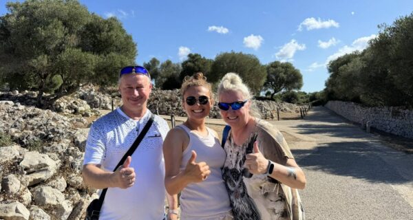 Troje uśmiechniętych turystów pozuje do zdjęcia podczas wycieczki na Minorce, pokazując kciuki w górę. W tle kamienne mury, ścieżka oraz drzewa oliwne.